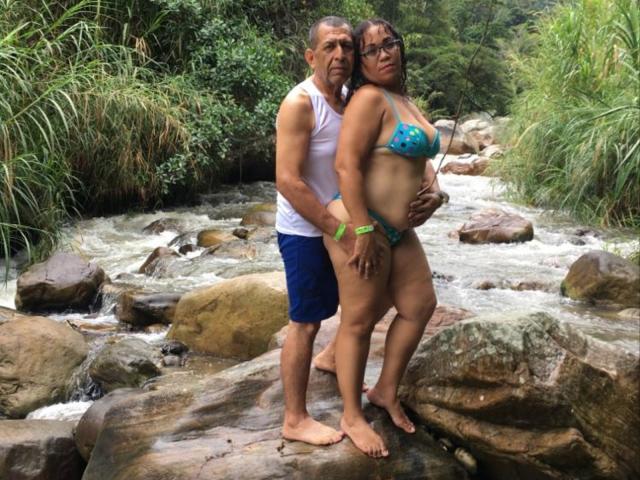 Pamela e Diego, usando um biquíni azul com seios grandes, estão em pedras, com os braços ao redor de um homem musculoso que veste uma regata branca e shorts azuis, em um riacho em uma floresta..
