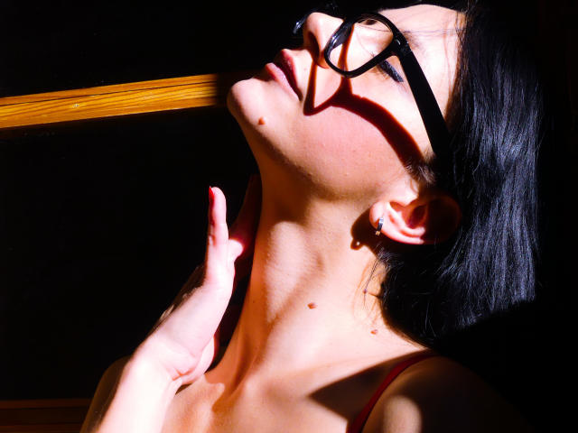 Cassandra Michelli con gafas negras, ojos cerrados, cabeza inclinada hacia atrás, luz solar en el rostro, mano cerca del cuello, lunares visibles, cabello oscuro, correa roja, fondo en sombra, piel iluminada por el sol..