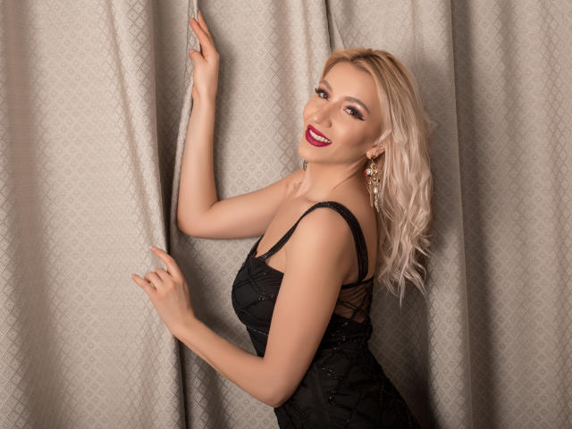 CyraVelvet, avec ses cheveux blonds, son rouge à lèvres rouge, sa robe en dentelle noire, tenant des rideaux, souriante, montrant ses seins, avec des boucles d'oreilles, bras droit levé, main gauche touchant le rideau, sexy, attirante..
