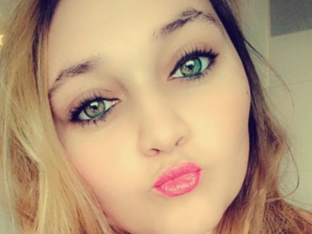 Sandyj a les yeux verts, des lèvres pleines roses, et des cheveux ondulés châtain clair, avec un trait d'eye-liner subtil et de longs cils. Son visage est légèrement incliné, et elle regarde l'appareil photo..