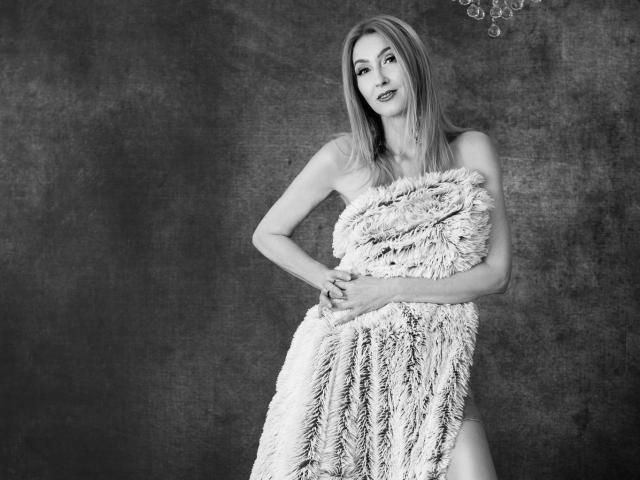 LadyLea with long blonde hair, standing, holding a fluffy, furry, white towel covering her tits and pussy, hands near her ass, in a black-and-white photo.
