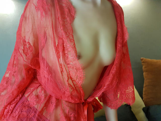 EroticaXEva veste um roupão vermelho de renda, expondo seus seios nus. O roupão é transparente, revelando sua pele clara e seus pequenos seios firmes. O roupão é amarrado na frente..