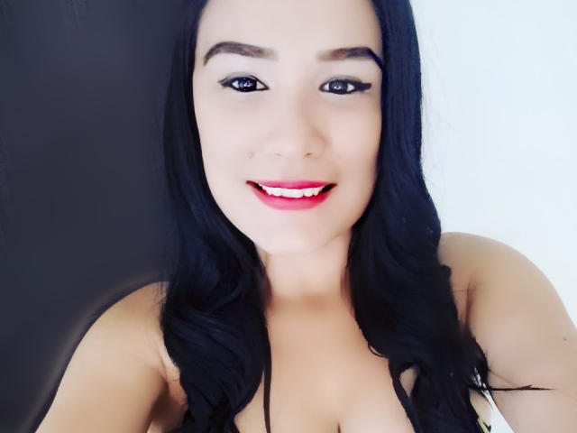 NathalieSexX, com cabelos pretos longos, batom vermelho e delineador preto, sorrindo para a câmera. Ela está mostrando os seios e tem um rosto bonito e sexy. Sua pele é clara..