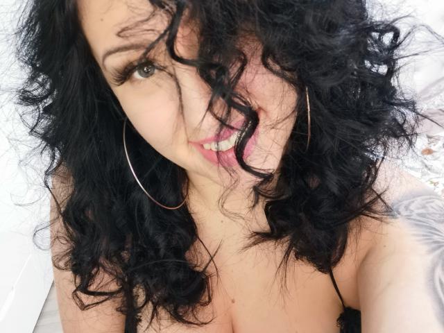 A LovelyMichellex69, com cabelos pretos cacheados, brincos de argola dourados e a língua de fora, exibindo seus seios em uma selfie, tem uma tatuagem no braço direito..