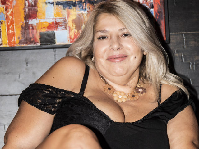 ByaStorm with blonde hair, black lace top, and beaded necklace, showing her big tits and smiling slightly. She's sitting with knees up, looking cute and confident.