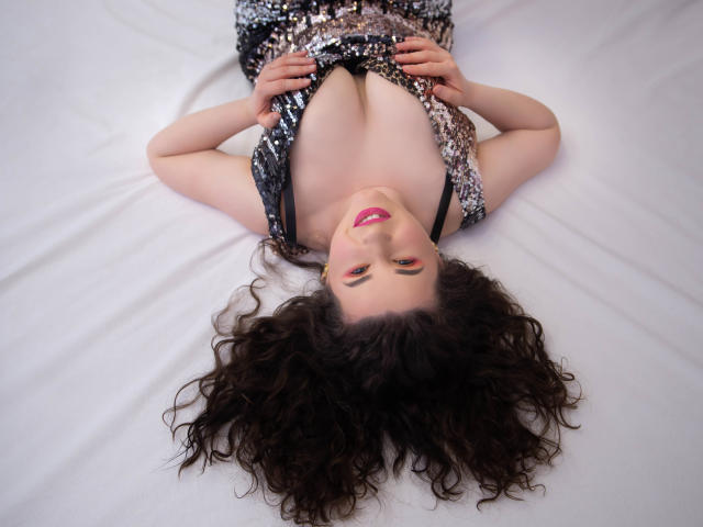 GorJesHot, con cabello castaño rizado, acostada sobre blanco, vestida con un vestido con lentejuelas, con las manos sobre el pecho, mostrando sus pechos, sonriendo con lápiz labial rosa, mirando hacia arriba..