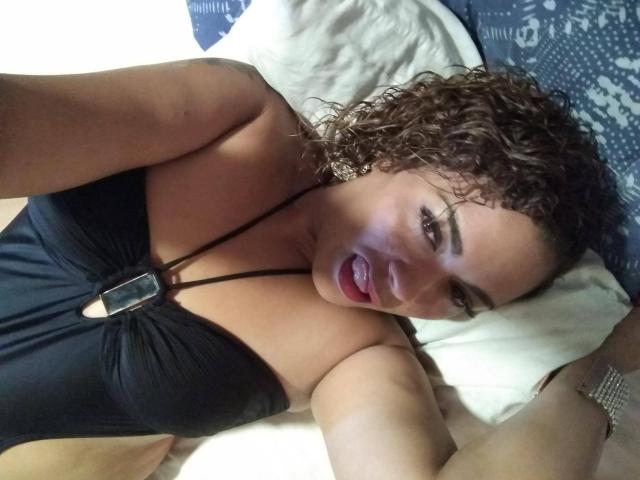 Solmary: Bruinharig, met krullen, draagt een zwarte haltertop, ligt op een bed, heeft haar tong uitgestoken, toont haar grote borsten en heeft een sexy glimlach, op een selfie..