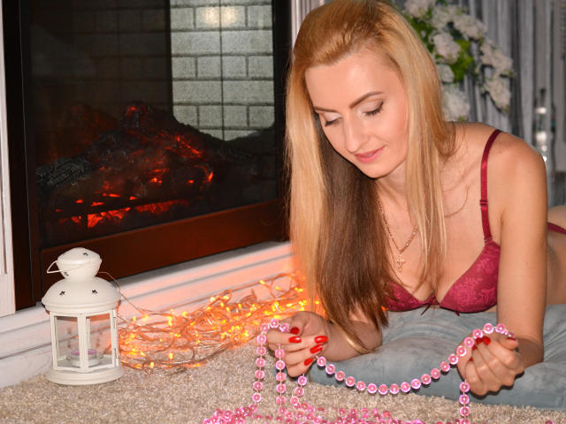 VotreRealGirl avec des cheveux blonds, un soutien-gorge rouge et des perles roses, tenant une guirlande de perles roses, appuyée sur un tapis près d'une cheminée allumée, portant un jean bleu, souriant doucement..