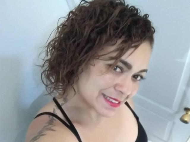 SolmaryHot with curly brown hair, smiling, pink lipstick, black tank top, tattoo on right shoulder, looking up, white door in background.
