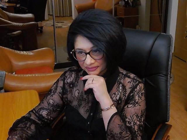 ClassybutNaughty with black lace top, glasses, and short black hair, sitting in a leather chair, resting chin on hand, smiling, showing red nail polish, and wearing a ring.