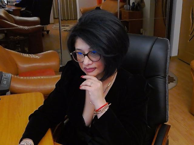 ClassybutNaughty with black hair, glasses, and red lipstick, wearing a black dress, sits in a leather chair, touching her chin with a ringed hand, smiling.