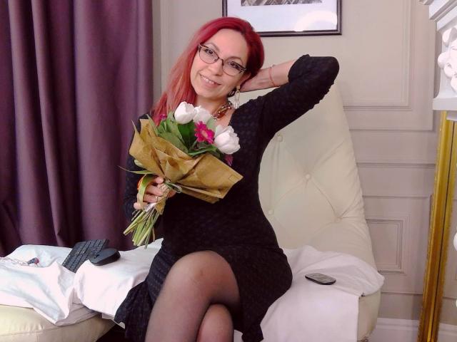 DirtyMature with red hair, glasses, black dress, holding white tulips, sitting on white chair, arm raised, smiling, legs crossed, black pantyhose, TV remote, pillow, curtain.