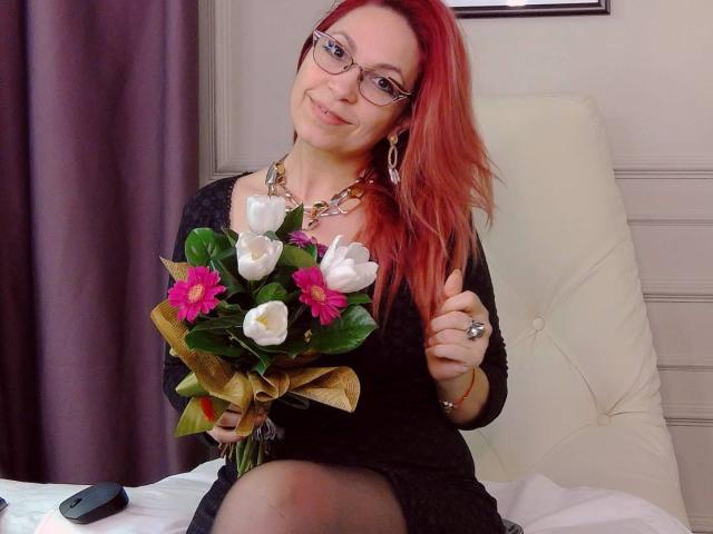 DirtyMature with red hair, glasses, black dress, holding white tulips with pink and purple flowers, gold necklace, and ring, sitting on white chair, smiling.