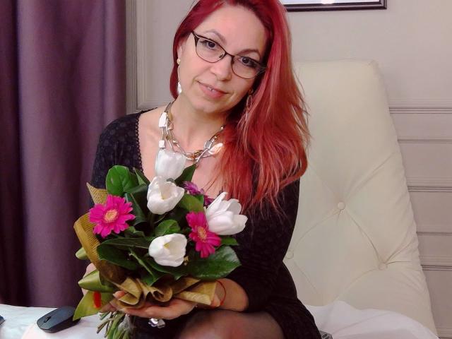 DirttyMature with red hair, glasses, and black dress, holding bouquet of white and pink flowers, smiling, wearing chunky necklace, sitting on white chair, looking cute.