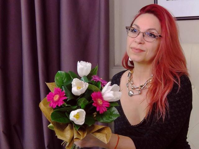 DirtyMature with red hair, glasses, and gold necklace holding a bouquet of white tulips and pink flowers, wearing a black top, smiling.