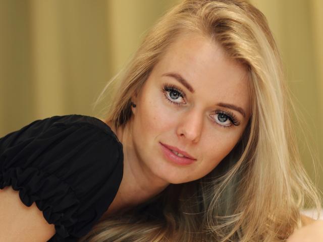ValeriexAngel with blonde hair, blue eyes, black shirt, and subtle makeup, looking directly at the camera with a slight smile, showing her soft, cute face and slightly parted lips.