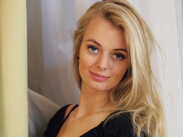 ValeriexAngel tem cabelo loiro, olhos verdes e um leve sorriso. Ela está vestindo uma blusa preta, com os seios discretamente visíveis. Ela é fofa e sexy, olhando diretamente para a câmera..