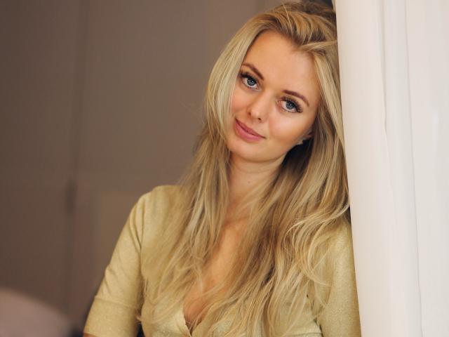 ValeriexAngel, com cabelo loiro, olhos azuis e um sorriso discreto, vestindo uma blusa dourada, apoiada em uma cortina branca, exibindo um rosto adorável e sensual..