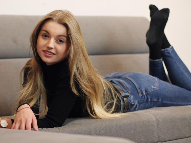 Erica, con cabello rubio largo, un suéter negro de cuello alto, jeans azules y calcetines negros, está acostada en un sofá gris, sonriendo, con las piernas levantadas, mostrando su trasero, y lleva un reloj de pulsera..