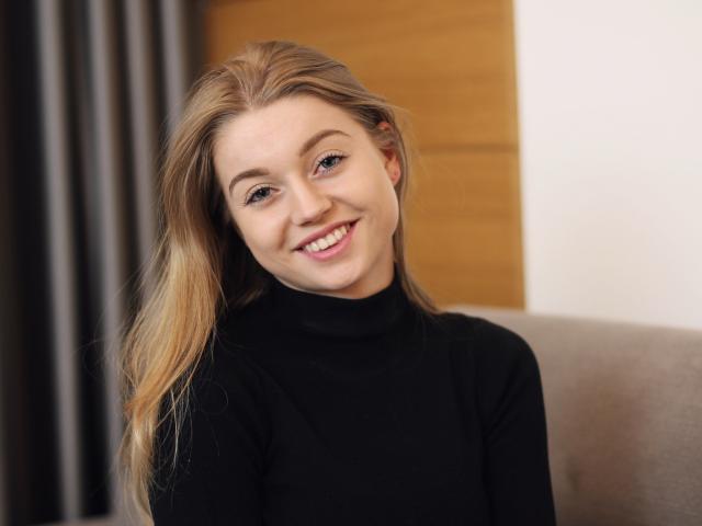 EricaLuck with long blonde hair, smiling, wearing a black turtleneck, sitting on a grey chair, soft light, slightly tilted head, relaxed, cute, natural makeup, gentle expression.