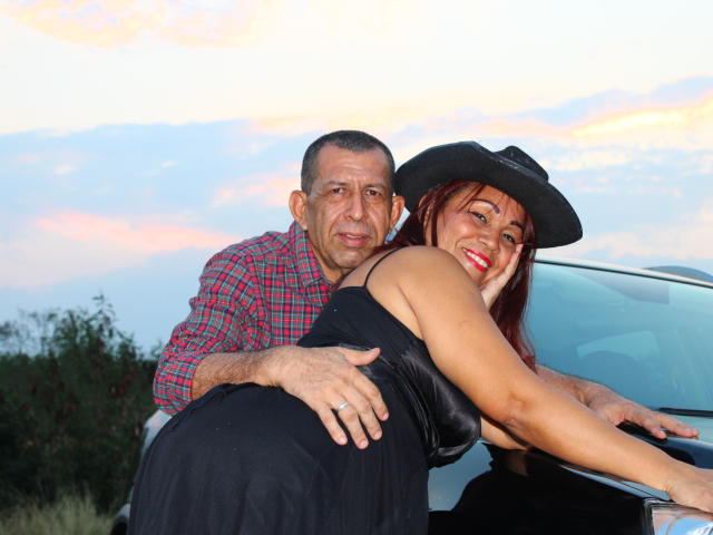 PamelaYDiego with tan skin, red lipstick, black dress, and hat, leaning on a car. Man in red plaid shirt, hand on her ass, smiling. Sunset sky background.