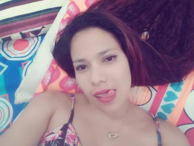 VenussHot with red hair, tongue out, looking up, wearing a floral bikini top, necklace with heart, lying on colorful patterned bed, light skin, subtle makeup.