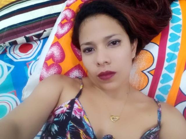 VenussHot lying on a colorful pillow, wearing a floral dress, with dark red lipstick, a gold heart necklace, and brown eyes, looking at the camera with a neutral expression.