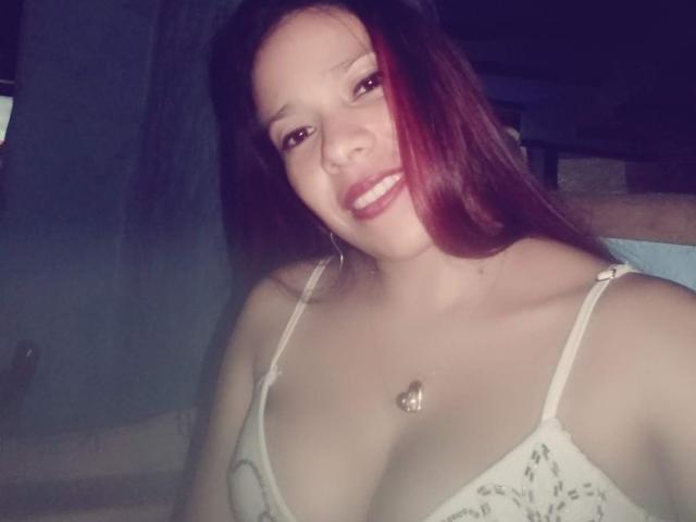 VenussHot with red and brown hair, smiling, wearing a white lace bra, showing her tits, has a heart necklace, and is in a dimly lit room.