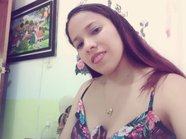 VenussHot has brown hair, a nose ring, and is wearing a floral dress with a heart necklace. She's smiling with red lipstick, showing her teeth.