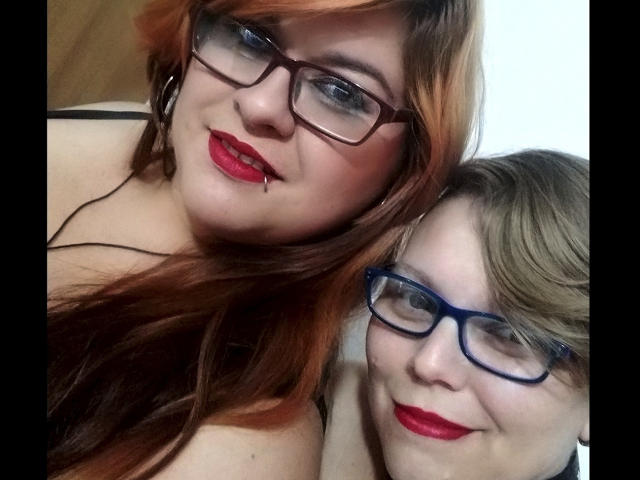 Twee Latina vrouwen met een stoere uitstraling, beiden met een bril. Eén heeft rode lippenstift en oranje haar, de andere heeft blauwe lippenstift en blond haar. Ze glimlachen dicht bij elkaar. Beide vrouwen hebben rode lippenstift en zijn topless..