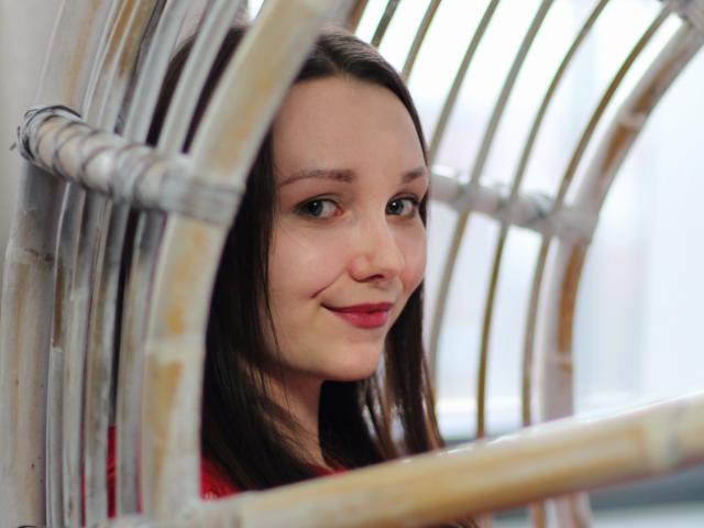 QueenZoe with brown hair, green eyes, and pink lips, smiling slightly, framed by wooden chair slats, wearing a red top, in a bright, softly lit indoor setting.
