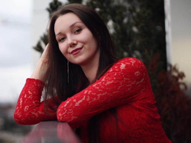 QueenZoe mit langen, braunen Haaren, einem roten Spitzenoberteil und einem leichten Lächeln, lehnt sich an eine Glasfläche und zeigt ihr süßes Gesicht und ihre kleinen Brüste, während sie lange Ohrringe trägt..