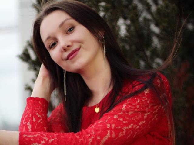 QueenZoe with long brown hair, wearing a red lace top, gold heart necklace, and silver earrings, smiling gently, her hand in her hair, showing a cute and sexy face.