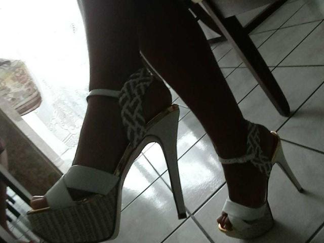 Kelcasada's legs in white high heels with silver chain straps, standing on shiny white tile floor. High heels are tall with gold soles and white platform.
