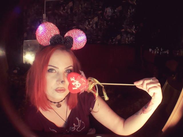 Uma mulher ruiva chamada ArdentBarb, usando orelhas brilhantes do Mickey Mouse, segurando um pirulito em formato de coração vermelho, com um piercing no nariz e uma tatuagem, vestindo uma camiseta preta, em um quarto com pouca luz..