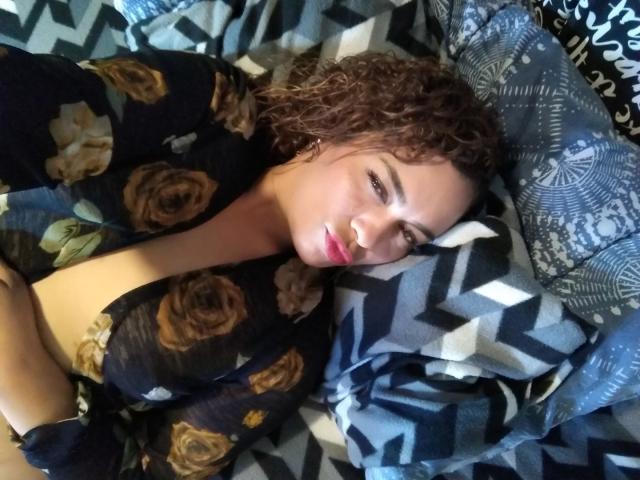 Solmary, les cheveux bruns bouclés, allongée sur un oreiller à motifs, portant une robe noire à fleurs, laissant apparaître ses seins nus, et regardant l'appareil photo avec un léger sourire..