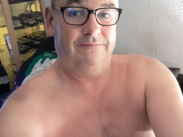 Davidoux sin camisa, con gafas, ojos azules y una ligera sonrisa, mostrando un poco de vello en el pecho y una pequeña barba de chivo. El fondo está borroso e indistinto..