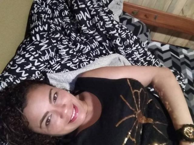 SolmaryHot with curly brown hair, smiling, wearing a black shirt with a gold spider design, lying on a bed with black and white patterned blankets.