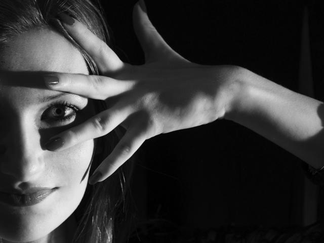 Fotografia in bianco e nero del viso di IampreciousLover, con una mano che copre un occhio, l'altro occhio che guarda intensamente in avanti, unghie dipinte di scuro, sfondo in ombra, tratti del viso delicati..