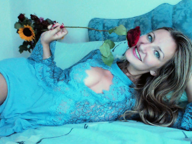 IampreciousLover está deitada em uma cama, sorrindo, segurando uma rosa vermelha, vestindo uma blusa azul de renda, mostrando seus seios, com girassóis ao fundo, um travesseiro azul atrás dela..
