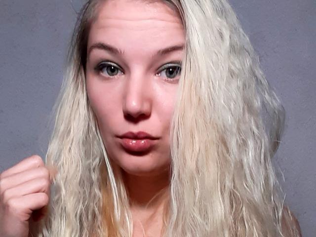 AlmaDeAngel69, con i capelli biondi mossi, la pelle chiara e le labbra rosa carnose, gli occhi leggermente socchiusi, il pugno alzato, guarda direttamente la fotocamera. Il suo seno è piccolo, la sua vulva non è visibile..