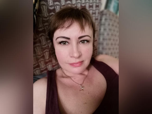 NanaGloss, avec ses courts cheveux bruns, ses yeux verts et son petit nez, porte un débardeur violet, un collier avec un pendentif en forme de clé, et sourit doucement à l'appareil photo..