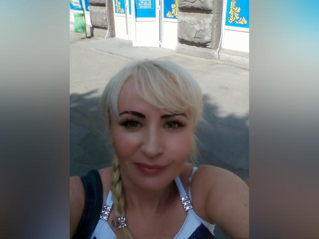 NanaGloss with blonde braids, wearing a white top with silver flower accents, has a cute smile, and is taking a selfie outside. Boobs are visible but not prominent.