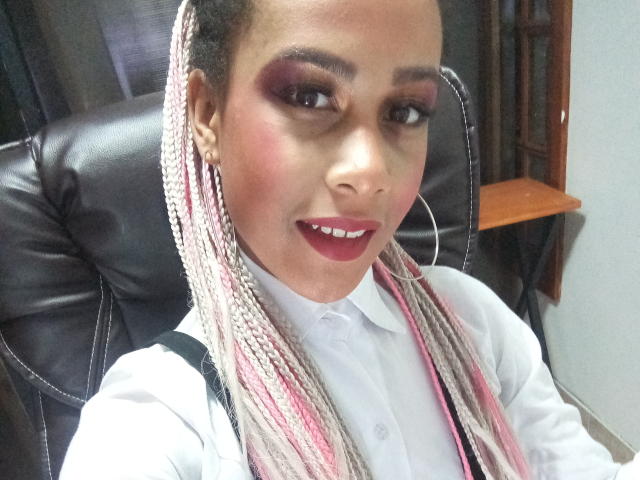 KendallQuinn with pink and white braids, dark red lipstick, white shirt, and hoop earrings, smiling at the camera, sitting in a black leather chair.