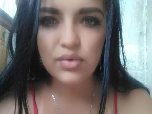 GavinGray tiene el cabello azul oscuro, cejas gruesas, pestañas largas y labios rosados. Lleva una blusa roja y un collar de plata, mirando directamente a la cámara..