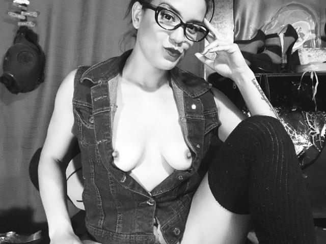 Schwarz-weißes Foto von ValeryGirlLatin mit Brille, einer offenen Jeansweste, die ihre Brüste zeigt, Kniestrümpfen, wobei ihre rechte Hand ihr Gesicht berührt, einem selbstbewussten Lächeln und einer Zigarette in ihrer linken Hand..