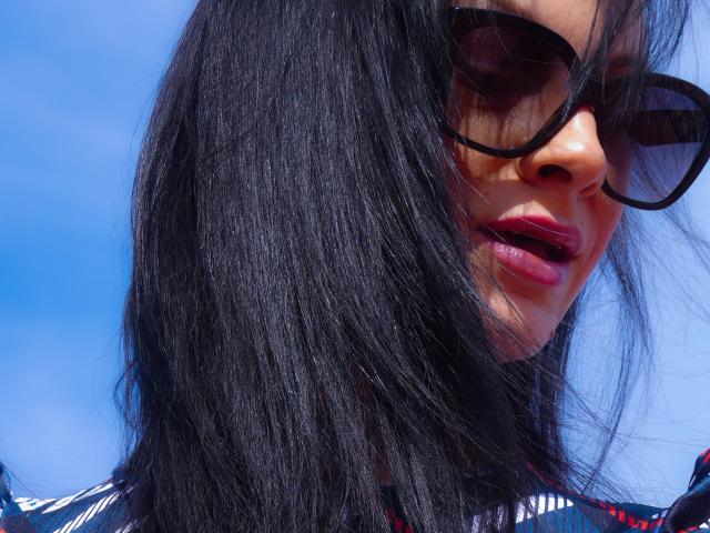 CassandraMichelli's face with black hair, dark sunglasses, and pink lipstick, showing a slight open-mouth smile. Her skin is tan, and her hair is shiny and straight.