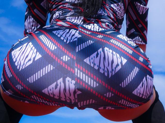 I glutei di CassandraMichelli, in un abito a righe zebrate, rosso e bianco, che mostra un tessuto aderente e lucido che mette in risalto il suo sedere rotondo e formoso. Sullo sfondo, un cielo azzurro..