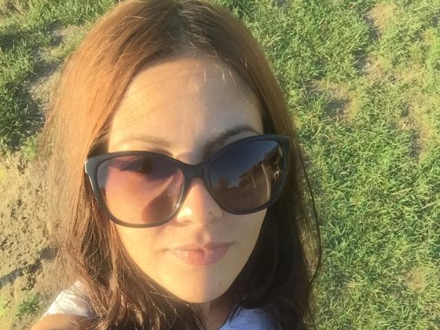 MyssJoy has brown hair, wearing black sunglasses, with light pink lips, and a slight smile, standing on green grass, sunlight on her face.