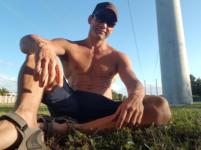 EdenAdonis ist ohne Hemd, sitzt auf dem Gras, trägt dunkle Shorts, eine Sonnenbrille und eine Mütze, hat eine muskulöse, gebräunte und verschwitzte Brust, und im Hintergrund ist ein schlaffer, unbeschnittener Penis zu sehen..